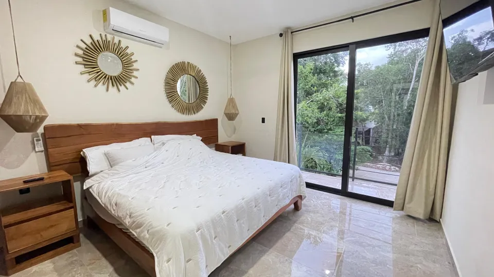 A bright bedroom with a wooden bed, a sun mirror and a glass door to the jungle.